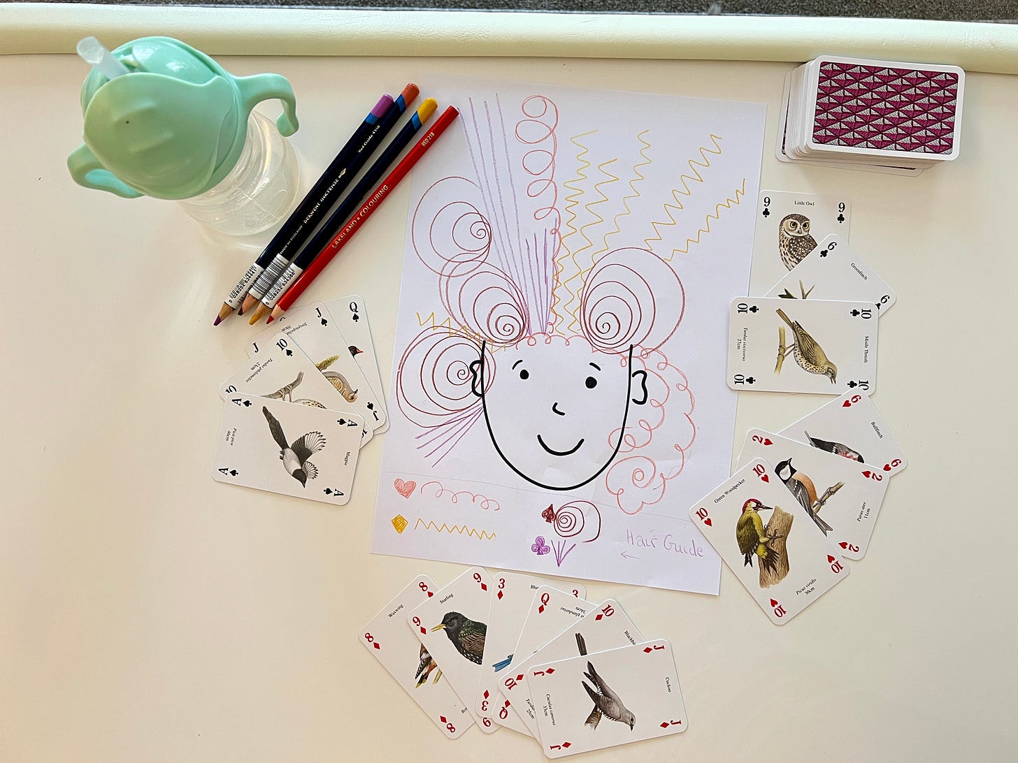 Hand-drawn cartoon face with hair made from different coloured pencil patterns. The patterns are linked to different playing card suits. The image is on a table and surrounded by playing cards, pencils and a child's drink bottle.