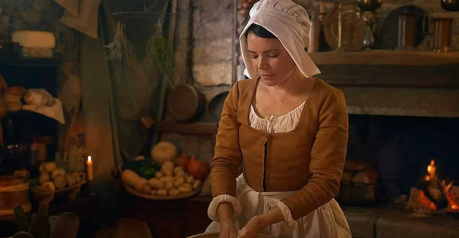 Imagem de uma atriz vestida como cozinheira no ano de 1663, em um cenário de época bastante decorado.