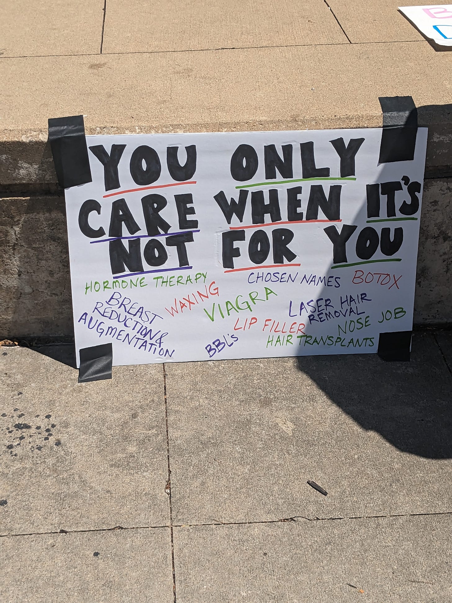 A photograph of a sign taped to the edge of a stage. It reads: "YOU ONLY CARE WHEN IT'S NOT FOR YOU. Hormone Therapy, Breast Reduction & Augmentation, Waxing, Viagra, BBL's, Lip Filler, Hair Transplants, Chosen Names, Botox, Laser Hair Removal, Nose Job"