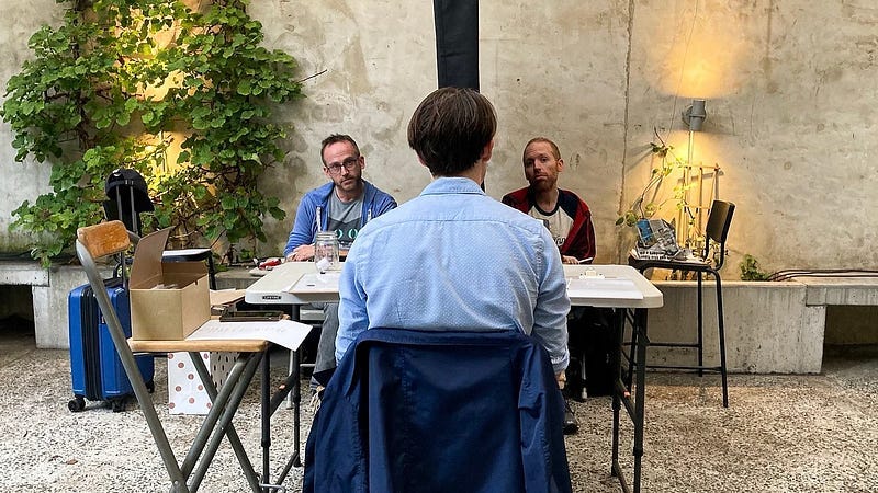 In the center foreground, a man sits facing away from camera and towards two other people. Those people are facing the man as they sit on opposite sides of a curtain, unable to see each other.