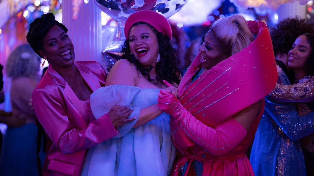 Three laughing Black women dressed up hug each other while dancing in a crowd amongst balloons at a party.