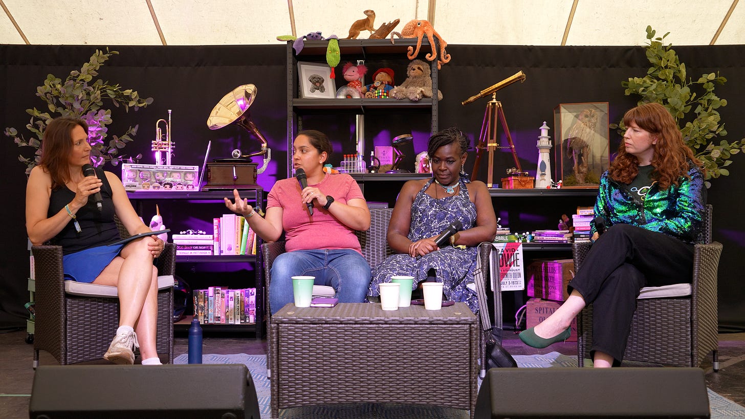 Four women sit on armchairs on a stage in front of shelves full of scientific equipment.