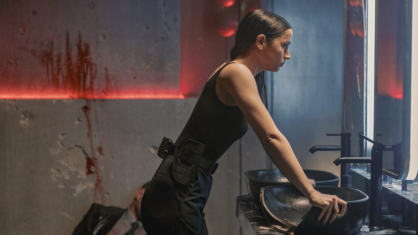 Eve braces herself over a shattered sink, bloodied and breathless, in front of a mirror in a neon-lit bathroom.