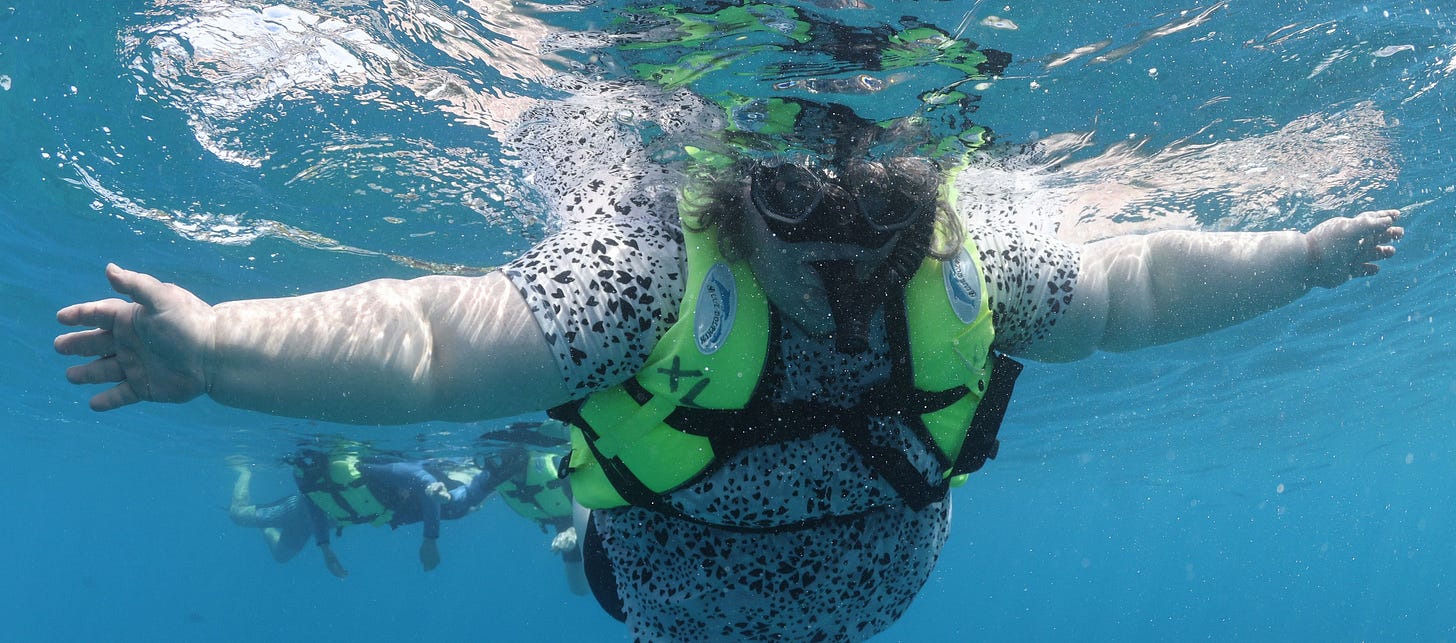 A fat woman floating underwater in mask and snorkel, arms outspread.