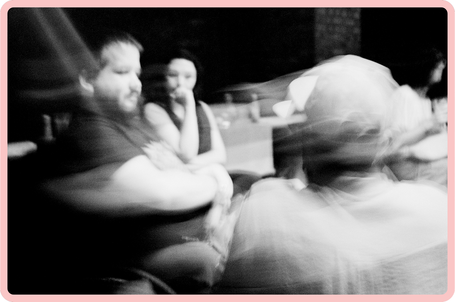 A black-and-white blurry photo of three young people talking. In the foreground we see the back of the head of a man with dark skin, and in the background, a bearded man who is a wheelchair-user and a young woman are sitting, listening.