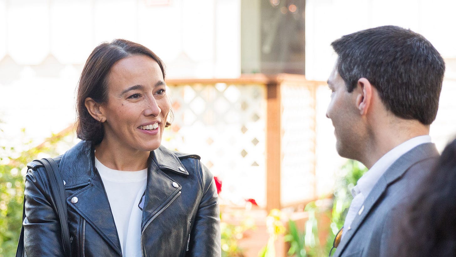 Two people are standing outdoors, engaged in conversation. The woman is wearing a black leather jacket, and the man is dressed in a light-colored suit jacket.