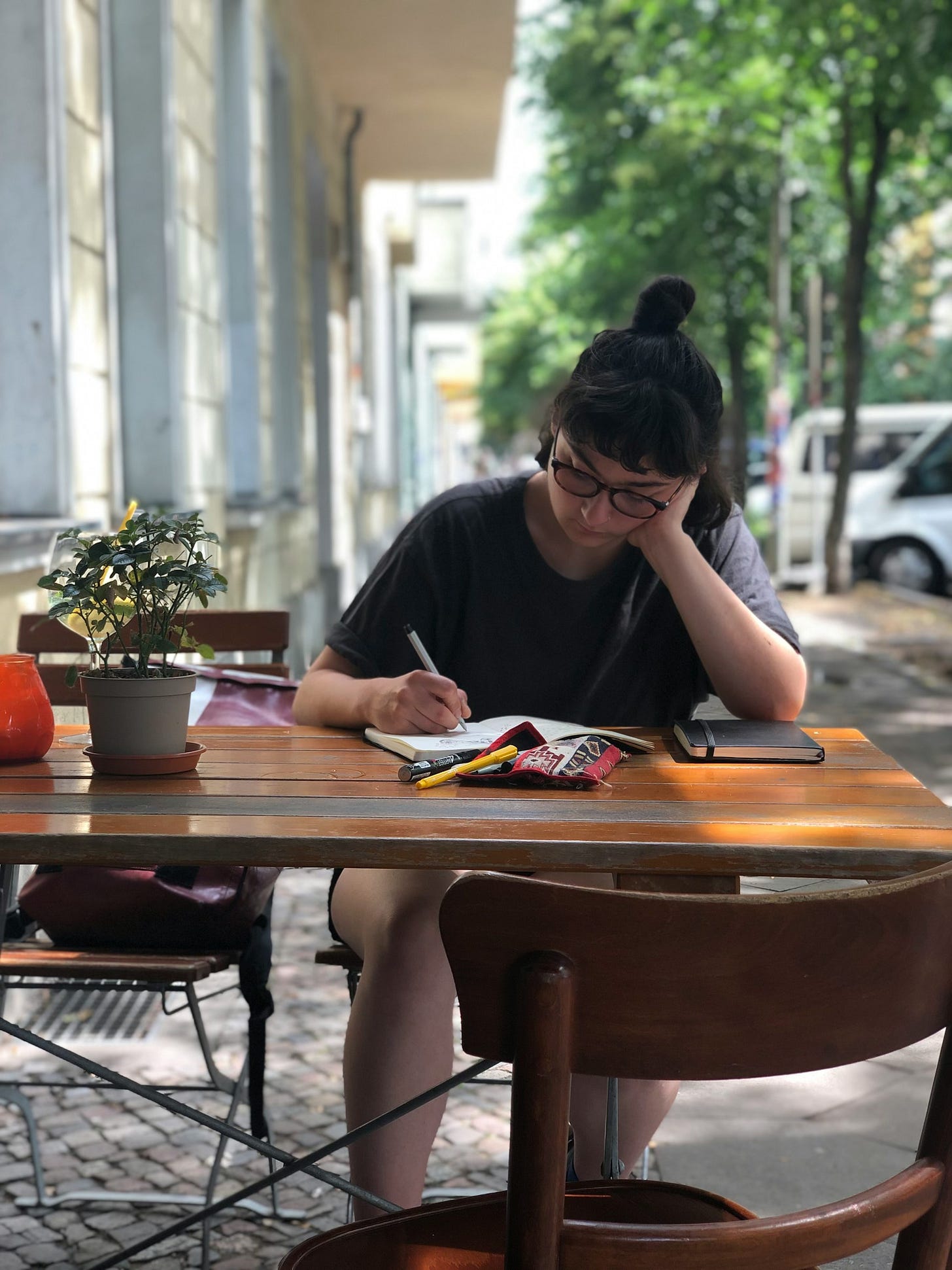 A woman wearing glasses, dark hair pulled back on top of her head, sits at an outdoor cafe table on a sidewalk, bent over a notebook, writing.