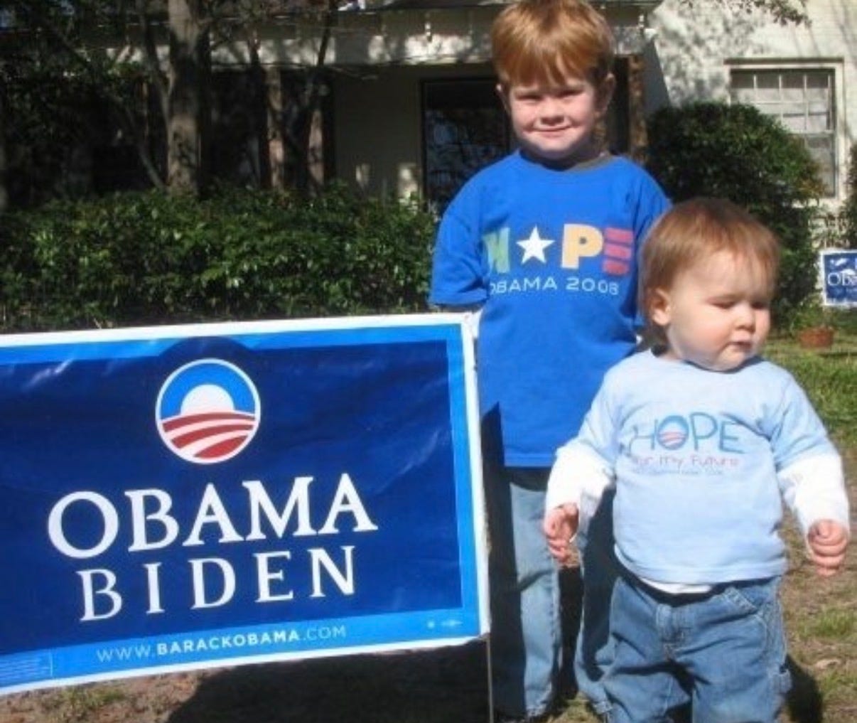 Two young boys standing on a lawn beside an Obama Biden sign