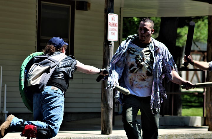4 cowled and masked figures at night holding a finger over their mouth shushing the camera. A LARPer striking someone dressed as a zombie, exposed ribs and gorey makeup, with a foam sword. LARPers standing in the woods. One wears a torn lab coat and reinforced leather armor, and another in an orange baseball cap weilds a nerf gun and has too many tool belts. That's me, with the orange baseball cap. A third is mostly out of frame, but their large foam axe sticks up into view. A collection of LARPers stand outside a cabin with "RH" on the wall and a pop-up tent outside. Weapons and gear lay around the area and on the table.