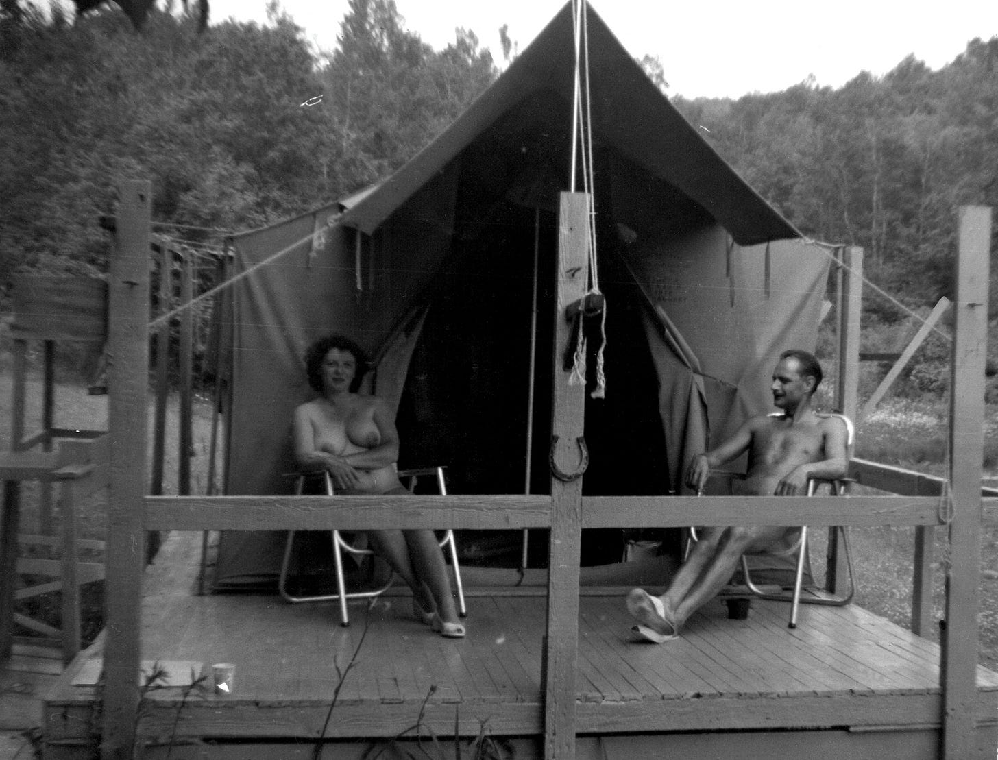 A black-and-white photo of two nude adults sitting in lawn chairs on the porch of a canvas tent cabin at a wooded nudist camp.