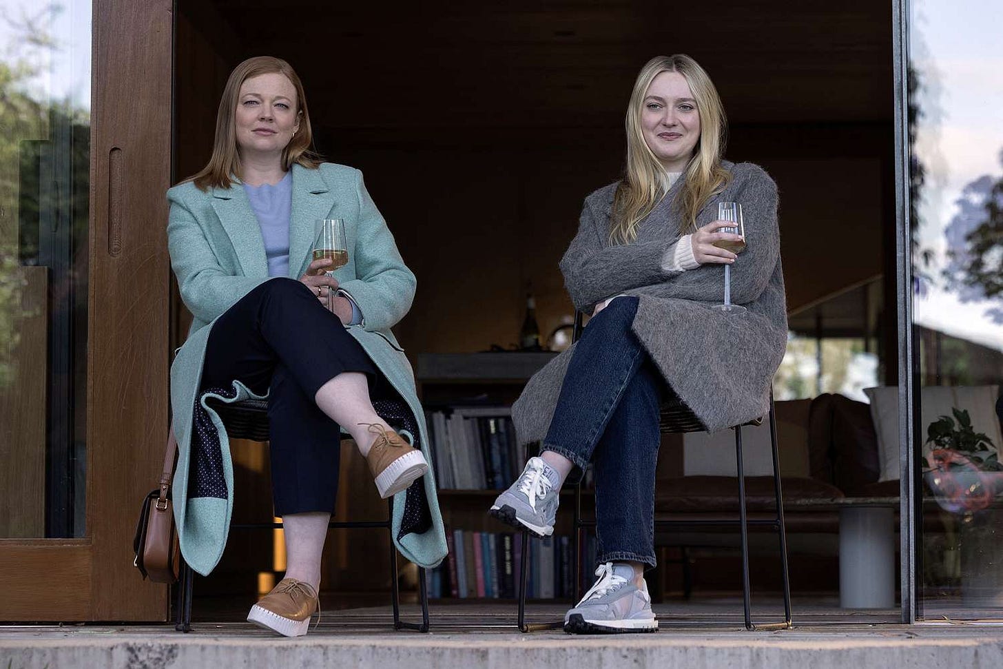 Two white women sip wine outside with their legs crossed towards one another, looking out and smiling at something off screen.