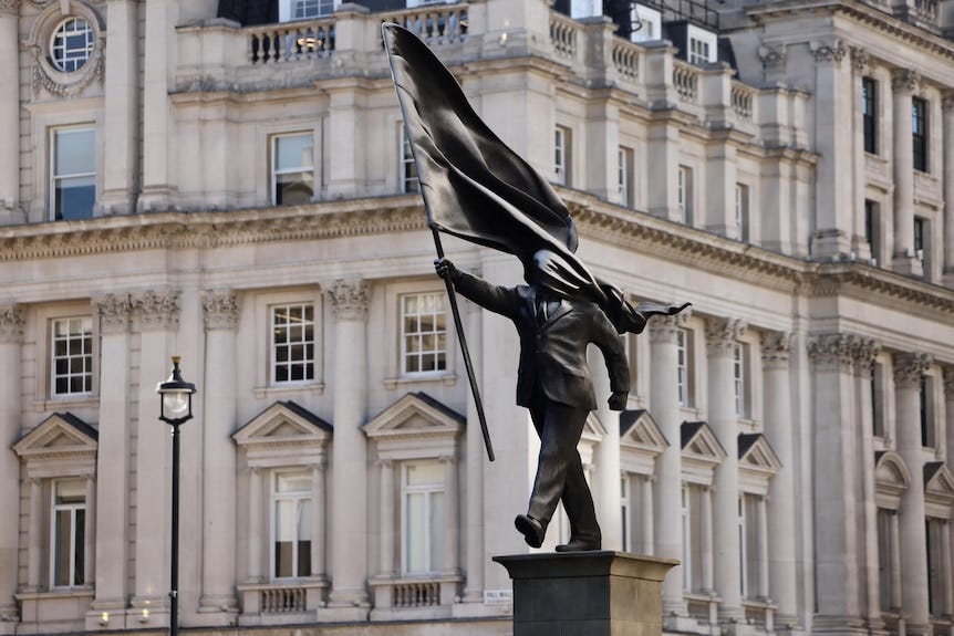 A statue of a man carrying a flag, blinded by it, about to walk off his statue plinth. A statue of a man carrying a flag, blinded by it, about to walk off his statue plinth.