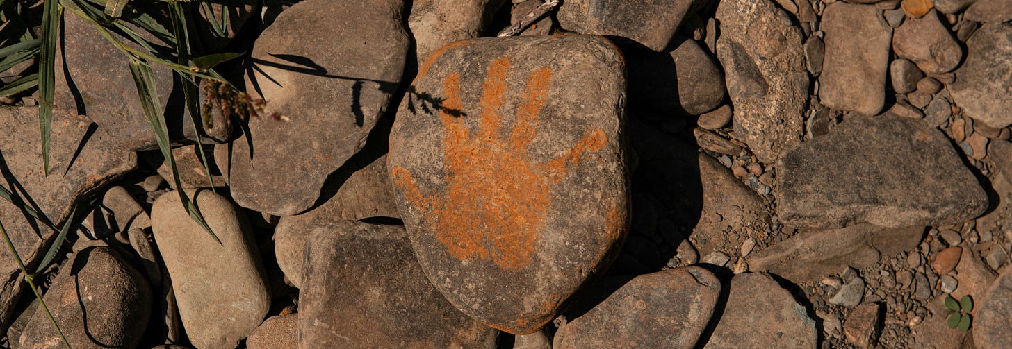 A weathered stone among river rocks marked with an ochre handprint, surrounded by dry grass — a quiet threshold between presence and passage
