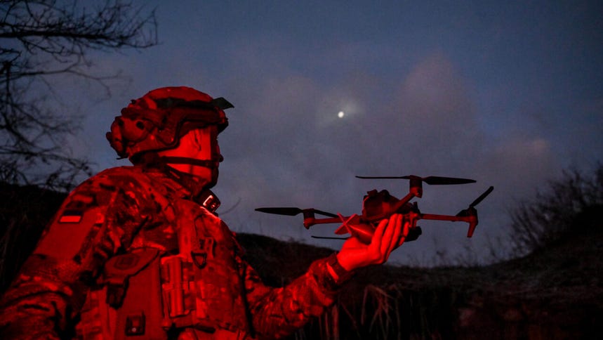 A soldier dressed in camo, bathed in red light, holds a drone at night.