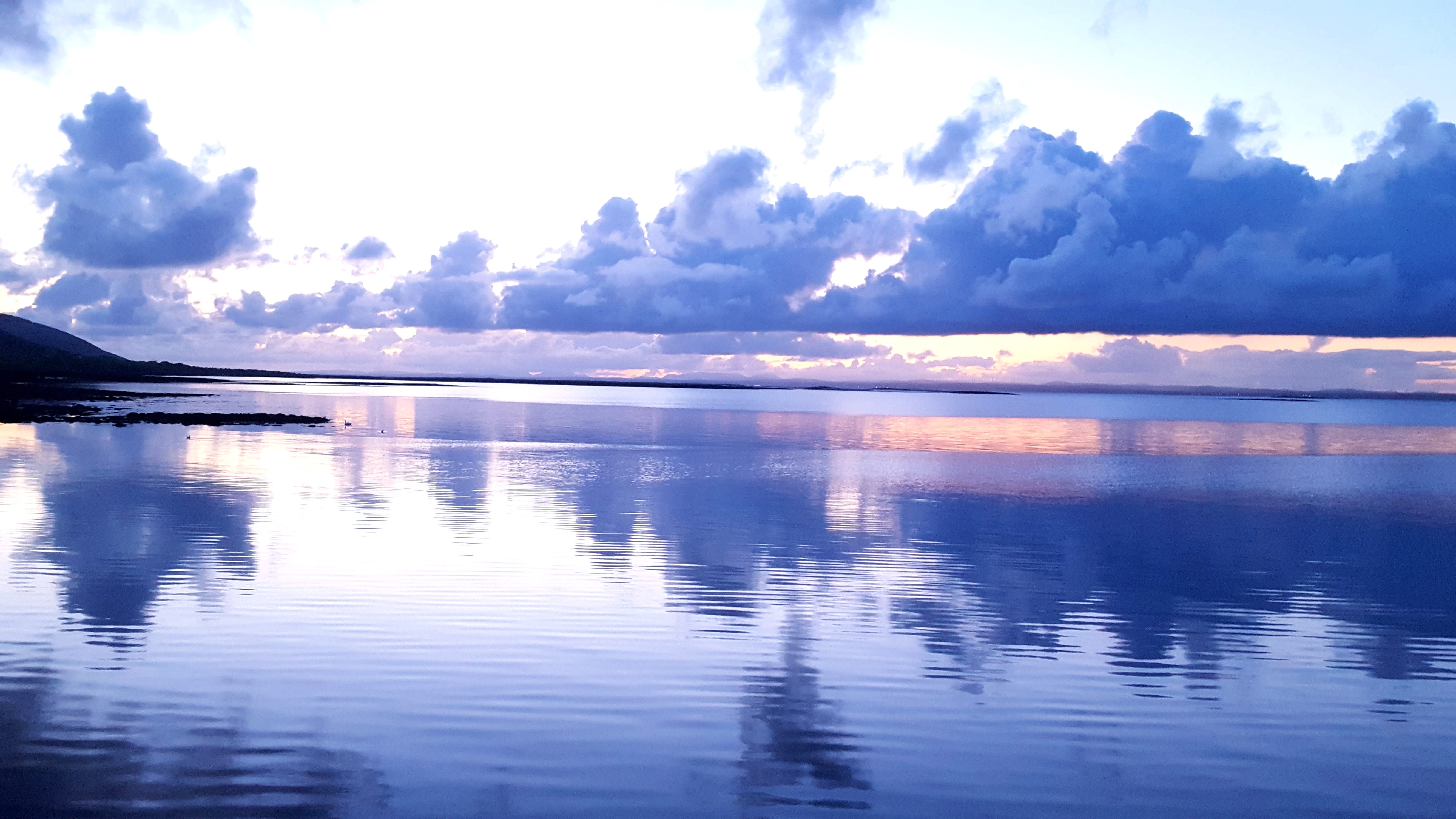 Looking out to sea at dusk, the tones of the image are lilac and blue due to the low light levels, the sea is flat and reflective like a mirror and there are towering banks of clouds in the sky.