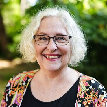 A wonderful photo of a friendly, smiling Shelley Rugg with a colorful print blouse and green trees behind her