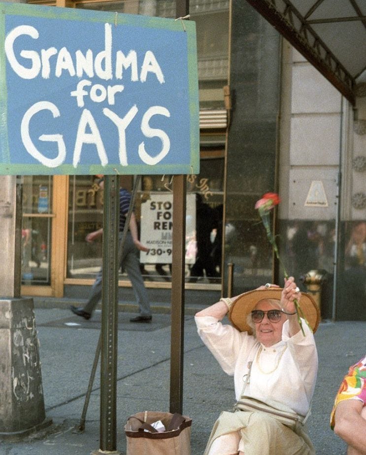 A grandma sits on a New York curb in a beige skirt, sensible heels, and a white button up. She holds onto her wide-brimmed hat while she waves one pink and one red carnation in the air next to a big blue and white, hand-painted sign that says, “Granda for GAYS”