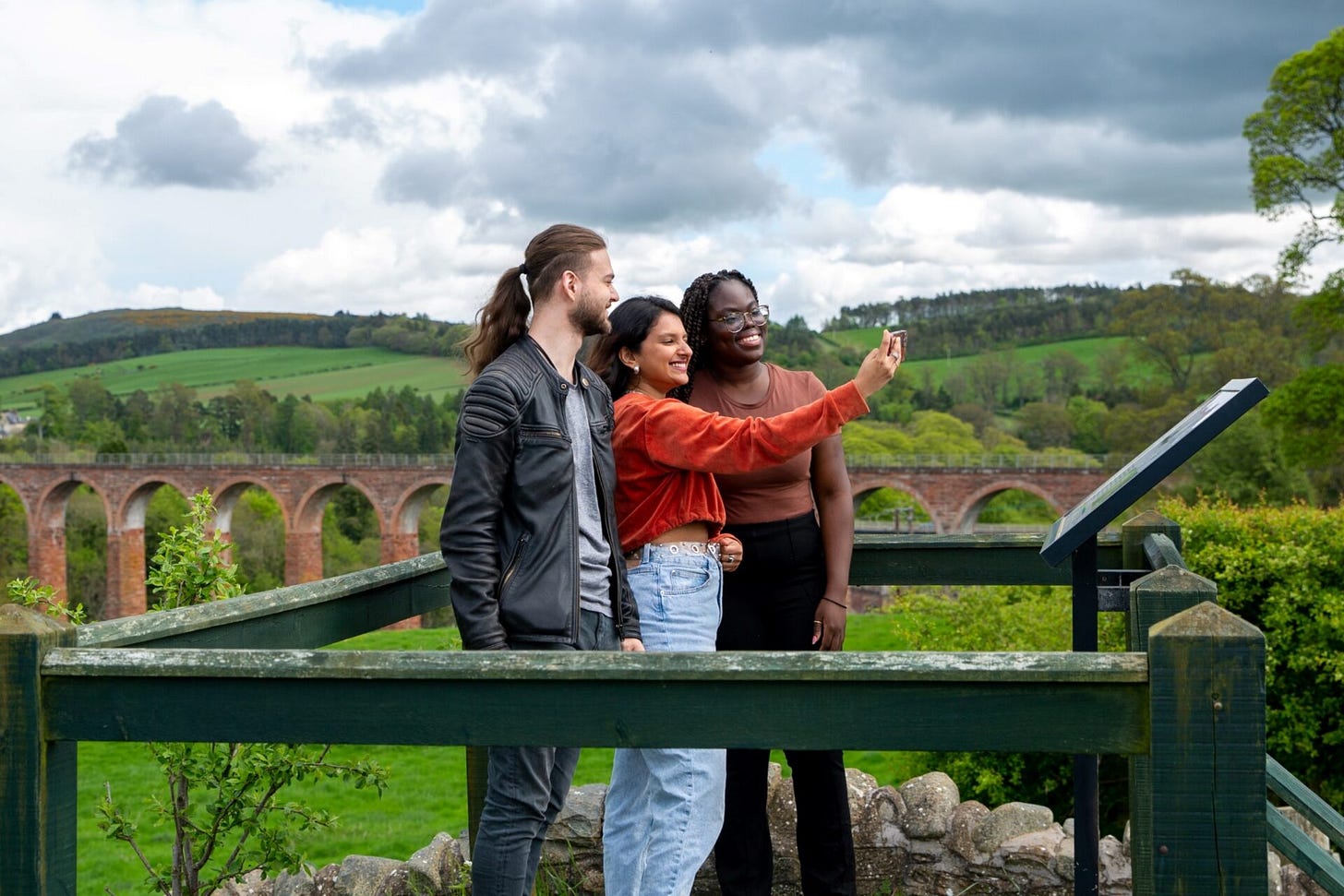 Three adults pose for a selfie in front of rolling hills, green trees, and a long red stone viaduct. The adult on the left has light skin, brown hair in a ponytail, and a beard. The adult in the middle has medium-tone skin and shoulder length dark brown hair. The adult on the right has dark skin, glasses, and long dark brown braided hair. Three adults pose for a selfie in front of rolling hills, green trees, and a long red stone viaduct. The adult on the left has light skin, brown hair in a ponytail, and a beard. The adult in the middle has medium-tone skin and shoulder length dark brown hair. The adult on the right has dark skin, glasses, and long dark brown braided hair.