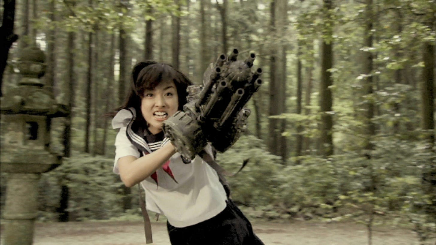 Woman in a schoolgirl outfit and a machine gun arm snarls and prepares to fire in a forest clearing