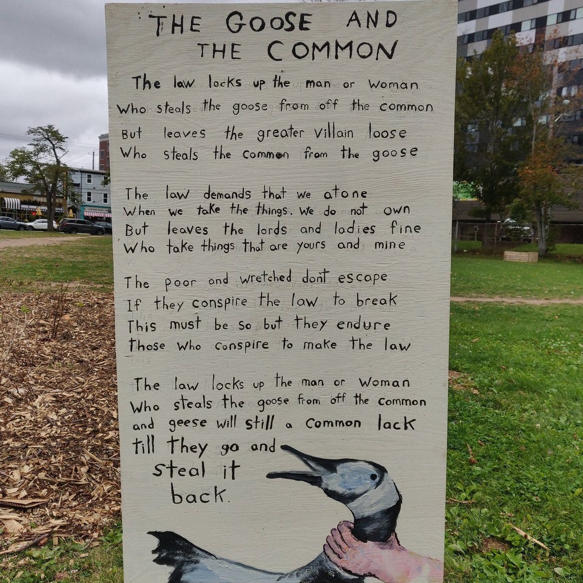 Handwritten poem titled The Goose and the Common on a signboard in an outdoor setting with grass, trees, buildings, and a car in the background. The poem discusses theft from the man or woman who steals the common from the goose, greater villainy, taking things not owned but leaving what is yours and mine, the poor and wretched must escape if they conspire but those who conspire to make the law endure, the law locks up the man or woman who steals the goose from off the common but lets the greater villain loose who steals the common from the goose. At the bottom, a drawing of a goose.