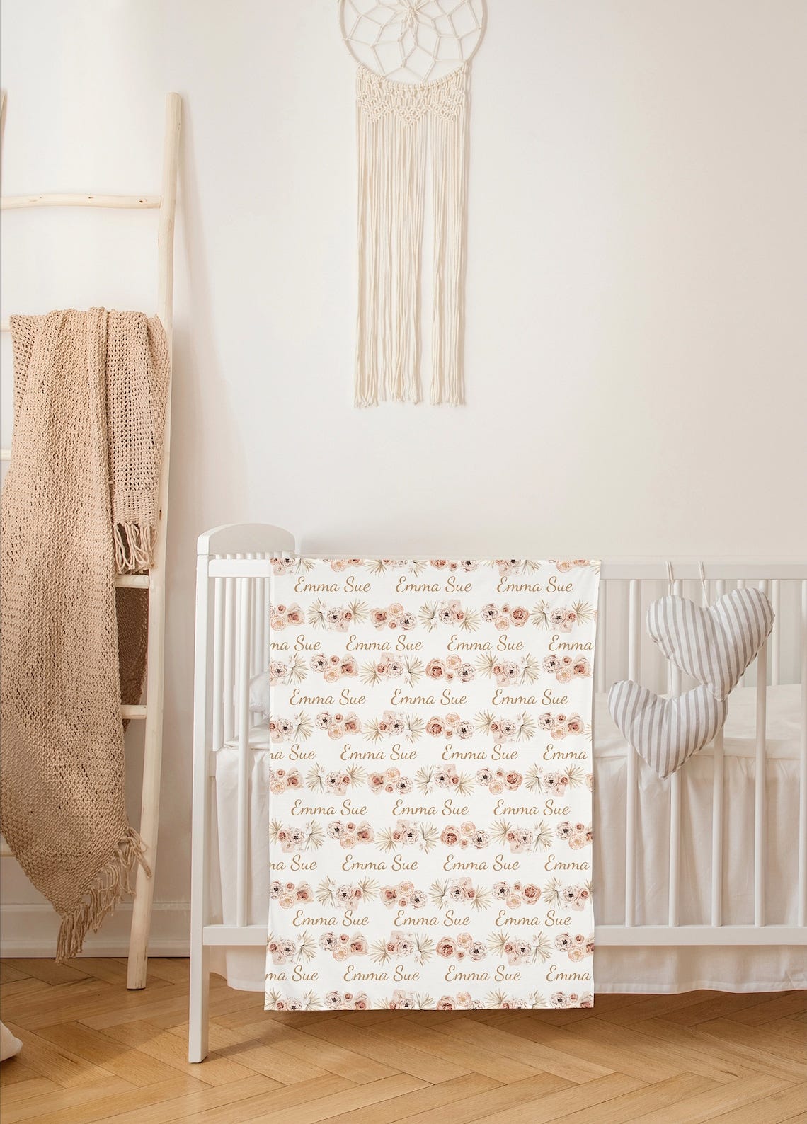 May include: A blush pink and cream baby blanket features a repeating pattern of watercolor flowers and the name 'Emma Sue'. The blanket is shown draped over a white crib in a nursery setting.  The design is soft and delicate, perfect for a baby girl's nursery. The floral pattern is subtle and elegant, and the name is tastefully incorporated into the design.