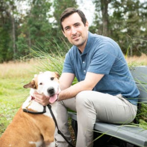 White man in blue short with dog. Background is trees and fields.