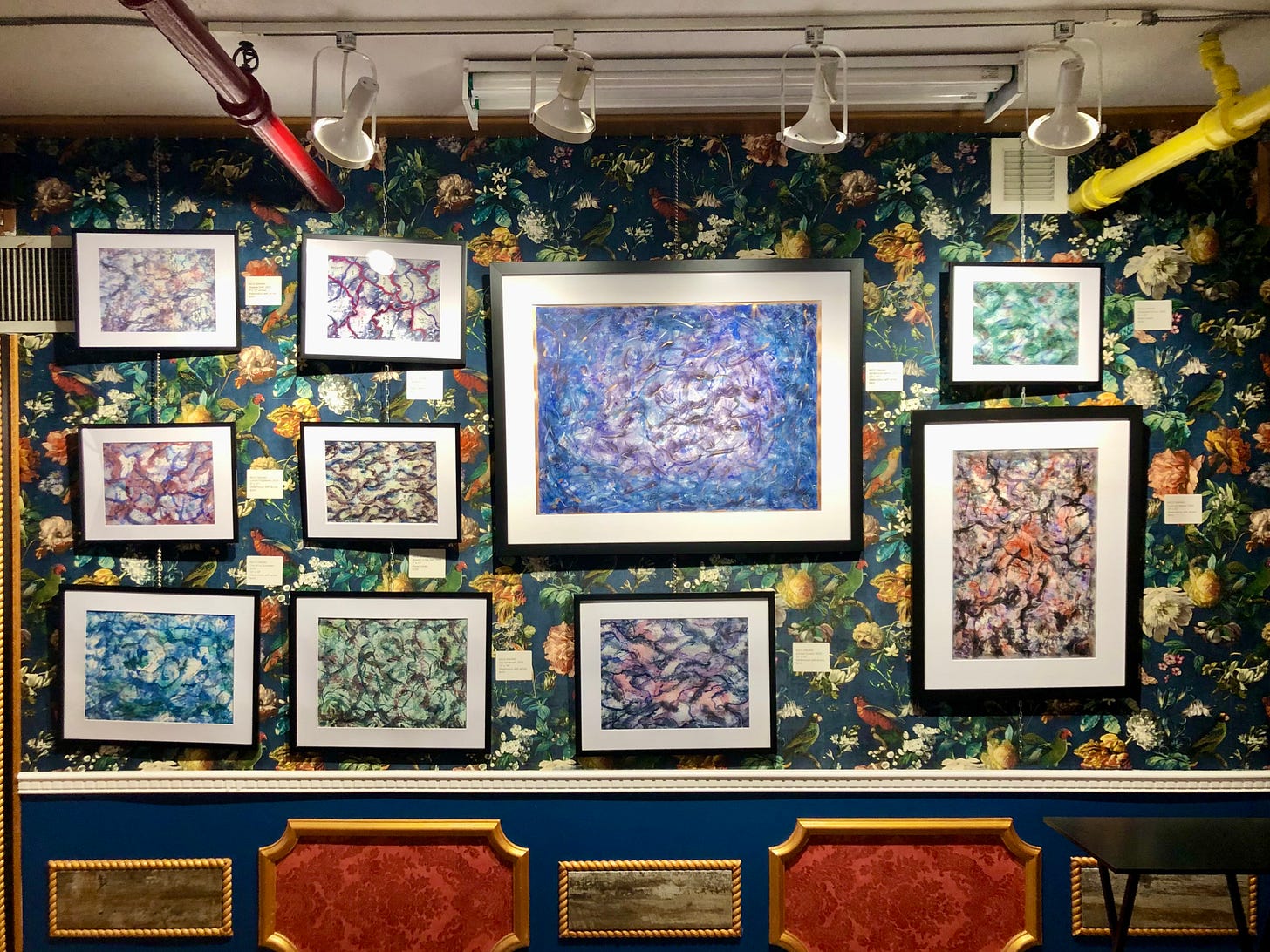 A frontal view of a richly decorated gallery wall filled with abstract paintings by Nico Sakaki. The artworks, framed in black and white, showcase dynamic, textured compositions with colors ranging from cool blues and greens to warm reds and purples. The dark floral wallpaper and ornate trim add a dramatic, vintage touch to the space.