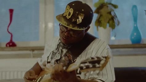 In first picture, Vumbi Dekula in a "BOSS" cap stares intently at his guitar as he plays it in on a couch in a sky-blue room. Second photo, the words "SLAVERY IS CRIME' is written in black outling over a scribbling of yellow, pink and black colors. In the third picture are four members of Dekula band.