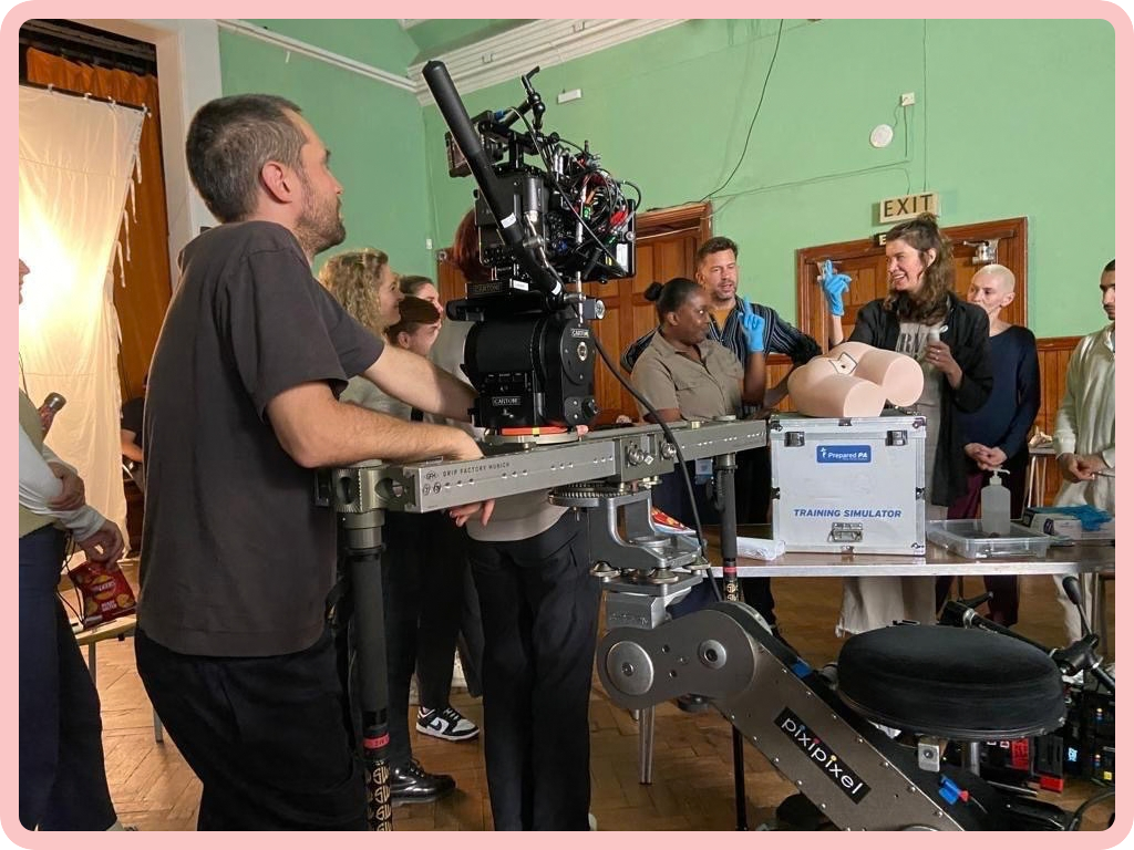 A photo of a film crew packed into a room with green-painted walls and large camera equipment. The cameras are pointed at a short black woman and a tall white woman, both of whom are wearing blue medical gloves and pointing an index finger upwards, while laughing. Beside them, on a large box, rests a plastic model of a human buttock, which gives the photo a sense of the absurd.