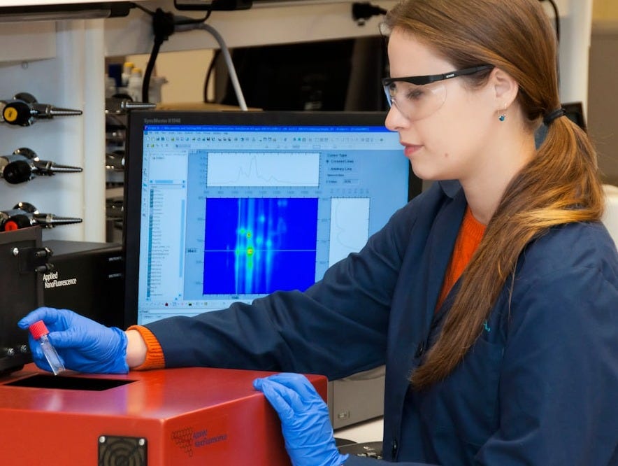 a female lab tech wearing a blue smock with black eyeglasses and holding a test tube