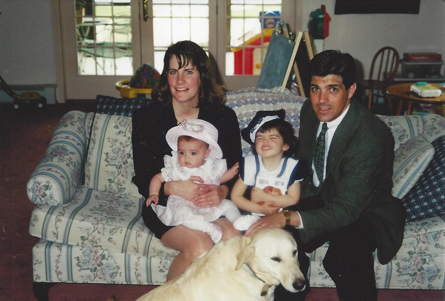 Husband and wife, a toddler girl, and baby girl--all dressed up for Easter--sitting on a formal sofa in a living room surrounded by toys and baby gear