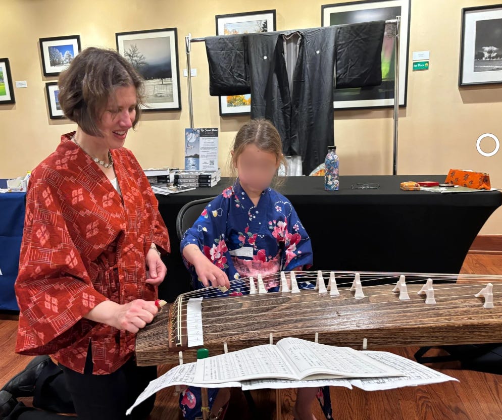 I, wearing a red Japanese top, coach a girl, wearing a blue yukata, or summer kimono, printed with hearts and blossoms, on how to play the koto. In the foreground is part of the koto, a wooden 13-stringed zither with movable bridges. I, wearing a red Japanese top, coach a girl, wearing a blue yukata, or summer kimono, printed with hearts and blossoms, on how to play the koto. In the foreground is part of the koto, a wooden 13-stringed zither with movable bridges.