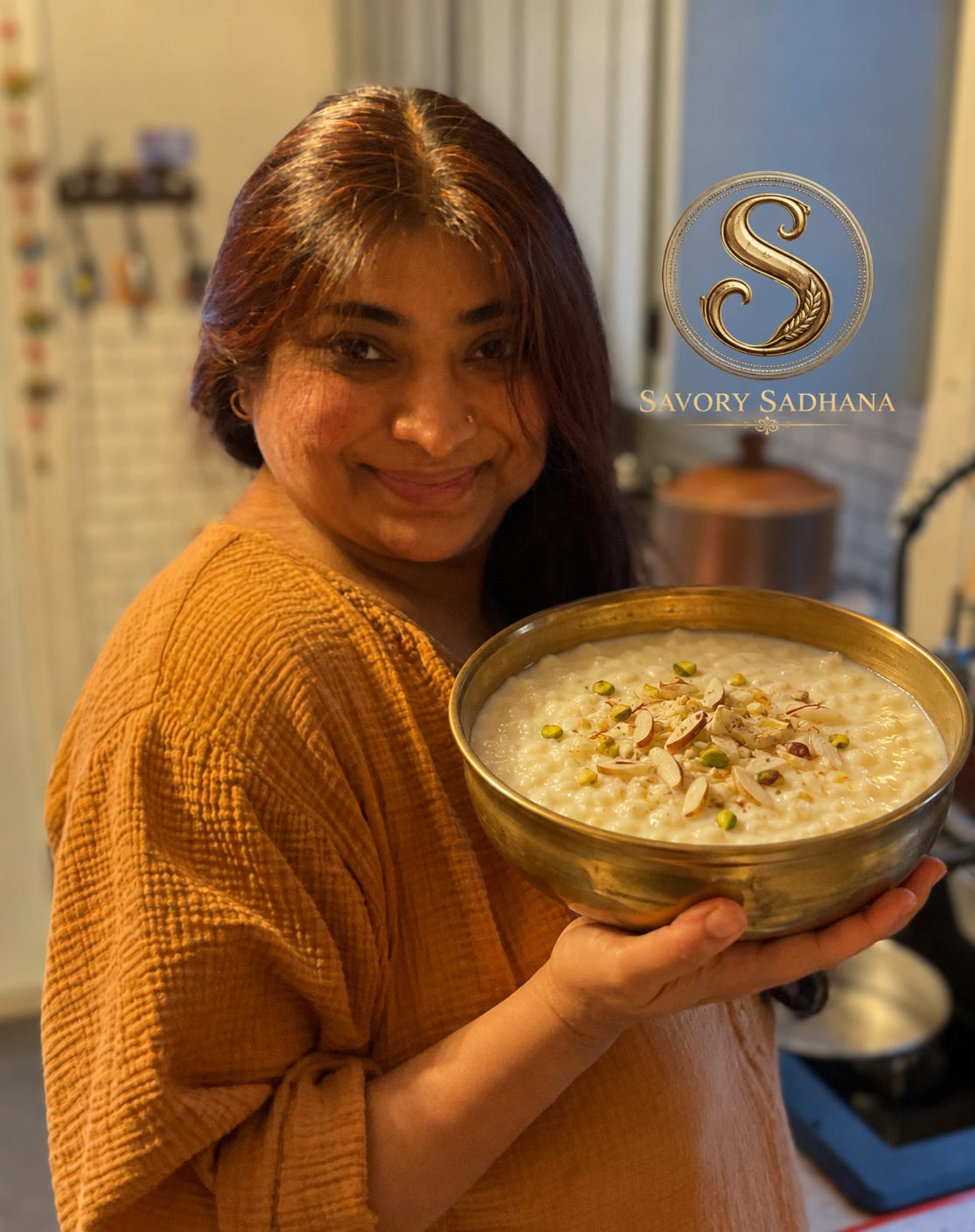Chef Daljeet B smiling in a softly lit kitchen while holding a brass bowl of traditional Indian kheer garnished with pistachios and almonds.