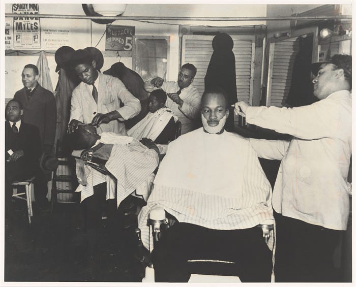 A black and white photograph of a barbershop on North Patrick and Pendleton Street in Alexandria, Virginia. The photograph features three barbers working on clients in the shop. Other clients wait in the background.