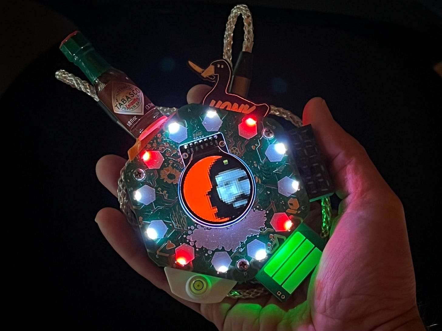 A small electronic device is being held in a hand and a bottle of Tabasco sauce has plugged into the side. The Documentally avatar is shown on a screen in its centre surrounded by red and white LEDs. There is also a duck holding a knife in its beak.