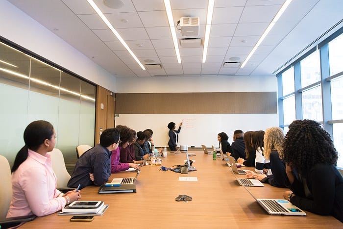 A woman presenting in the far background on a whiteboard, with a bunch of people sitting in chairs looking at her on both sides. A woman presenting in the far background on a whiteboard, with a bunch of people sitting in chairs looking at her on both sides.
