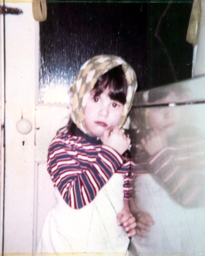 Young Lori at three years old, wearing a colorful patterned headscarf and striped shirt, looking directly at the camera with quiet curiosity — the photo that started everything.