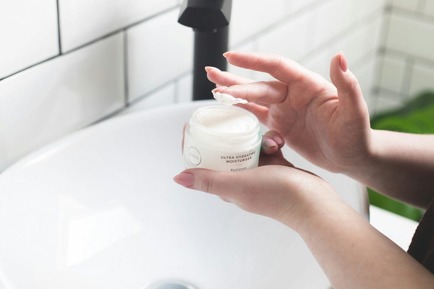 A woman holds a jar of cream in one hand while dipping her finger into it with the other.