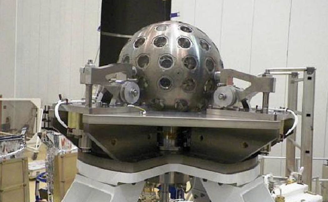 Image of a spherical silver object with regularly spaced features on its surface. It's he'd in a large room by a complicated support structure. Image of a spherical silver object with regularly spaced features on its surface. It's he'd in a large room by a complicated support structure.