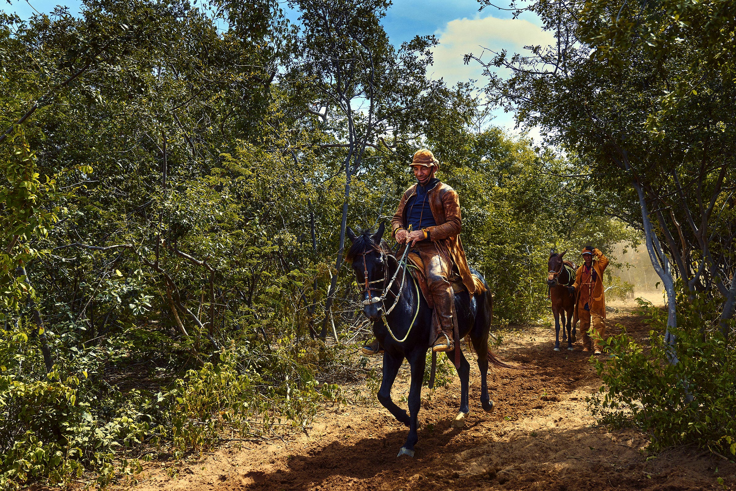 Three people ride horses along a dirt path through a lush, green forest. The leading rider wears a brown outfit and hat, followed by two more riders in similar attire amid sunlight and shadows.