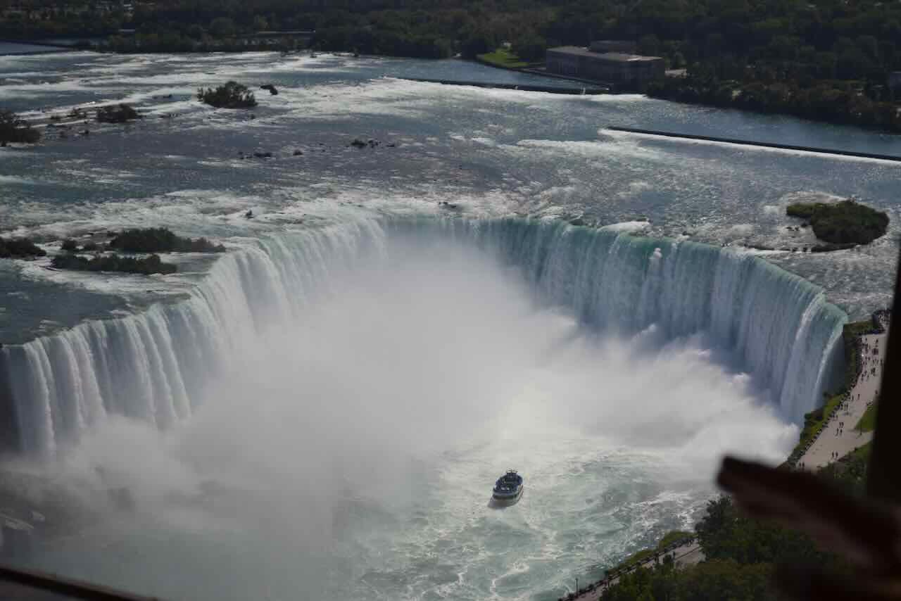 A tour boat approaches the base of Horseshoe Falls at Niagara Falls, surrounded by mist and cascading water, with lush greenery and observation areas visible along the river’s edge.