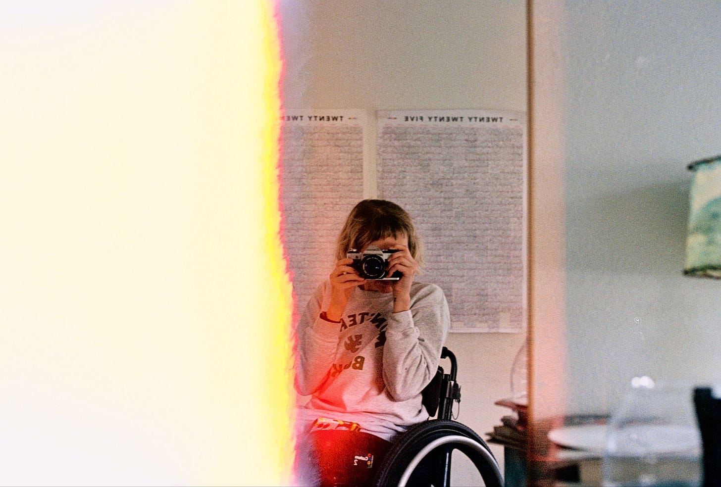The first photo on a roll of 35mm film, half the left side of the image is a blank half-burned frame. The edge is marked by a jagged yellow line that almost makes it look like a burning edge. The right side of the photo shows Rebekah taking a picture of herself in a mirror, her camera covering most of her face. She's sitting in her wheelchair wearing a sweatshirt that says "Monstera's Books." Three cheers for local bookstores!