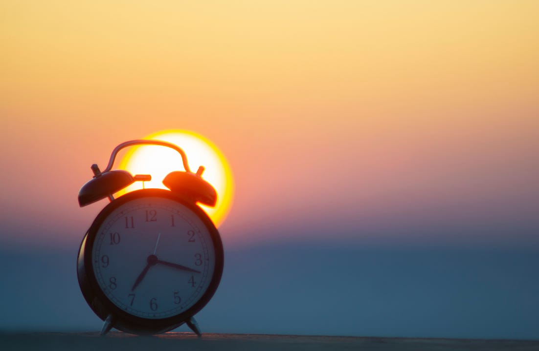 Analog clock face with a warm sunset in the background, soft orange light, calm and quiet mood. A visual metaphor for the biological timing shift at the center of the ADHD circadian clock.