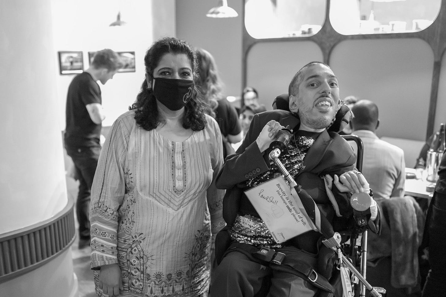 IMAGE DESCRIPTION: Within the Darjeeling Express restaurant, a black and White photo of Sulaiman (a wholehearted Disabled AF and British-Pakistani man with black buzzcut hair) sitting in his power wheelchair at the fully elevated height next to Asma Khan (a South Asian woman with black shoulder length hair), owner and head chef at the Darjeeling Express. Sulaiman is wearing his blazer with matching trousers, his sequinned shirt, and his pendent with the sign language symbol of “I Love You” (representing a universal meaning of love) via @RoseBYANDER. Asma is wearing a black face mask and a floral print kurta. Sulaiman is smiling and feeling joyful, having an inner fangirl moment in the presence of the legendary Asma. DESCRIPTION ENDS. 