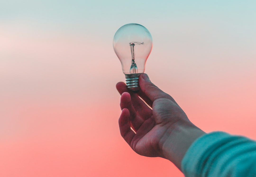 Outstretched arm, hand holding light bulb with a filament in it, against a pink and blue sky