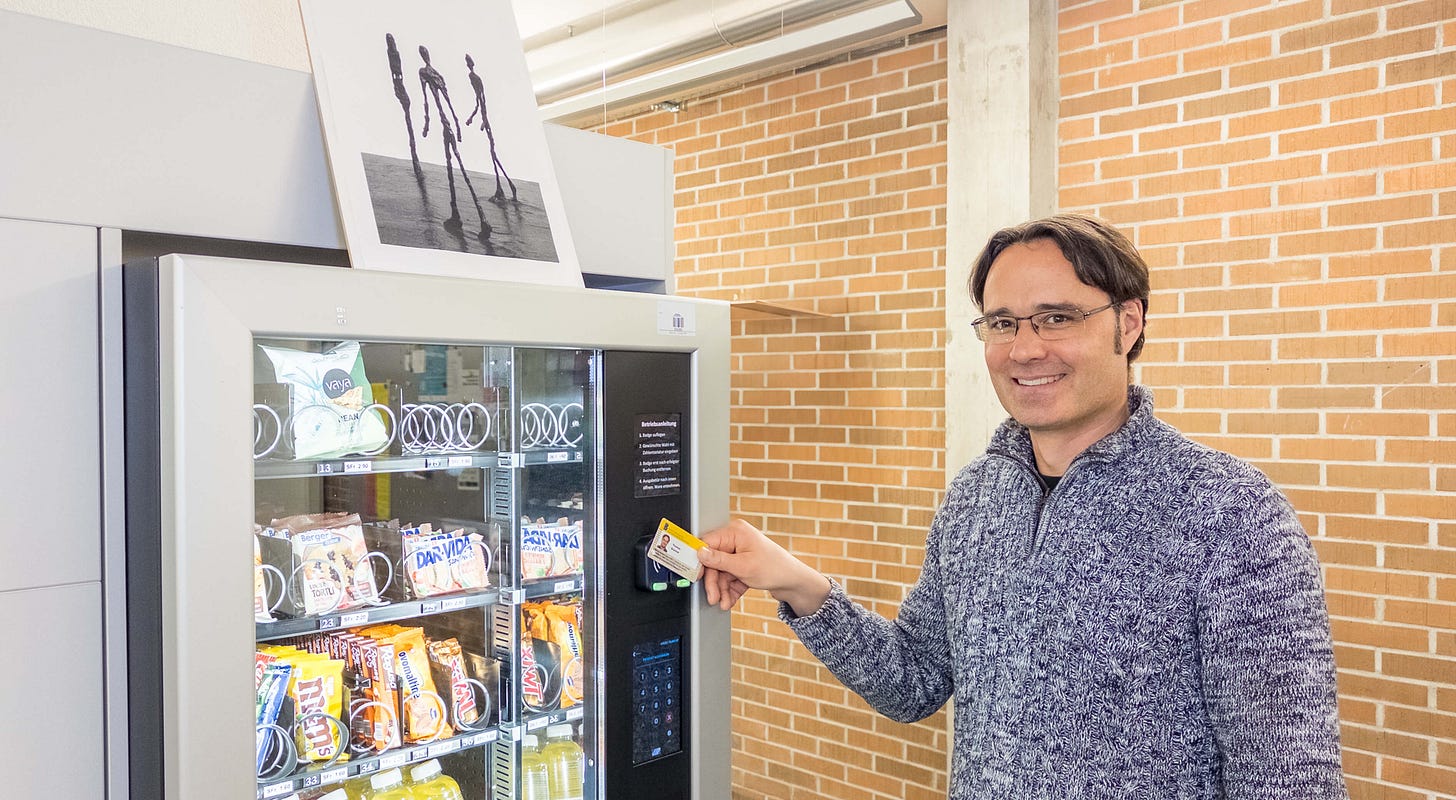 Das Foto zeigt den Psychologen Thomas Brunner, der lächelnd mit seiner Karte einen Snack aus einem Verkaufsautomaten holt. Der Automat bietet eine Auswahl an Snacks, darunter ungesunde Optionen wie Schokoriegel und Chips sowie gesündere Alternativen wie Nüsse und Müesliriegel. Auf dem Automaten steht ein Bild, das die berühmten, überschlanken Figuren von Alberto Giacometti zeigt. Der Hintergrund des Bildes ist eine Wand aus roten Backsteinen und eine helle, moderne Umgebung.