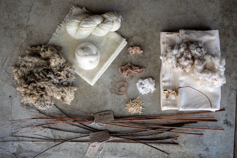 Raw undyed unbleached fabrics sit folded on a concrete floor, with piles of other fibres, yarns, woody stems and raw unprocessed wool.