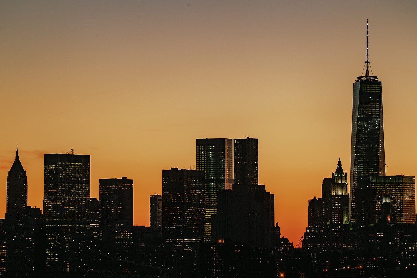 The Lower Manhattan skyline is silhouetted against a golden orange sky at sunset. One World Trade Center is prominent on the right, and small lights are visible in the windows of the dark buildings.