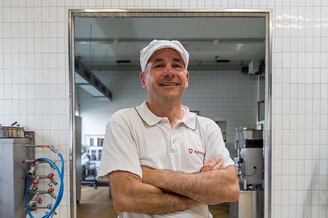 Eine Bildergalerie aus der Versuchskäserei von Agroscope zeigt verschiedene Schritte der Käseherstellung: Mitarbeitende in weissen Kitteln und Hauben im Gespräch, das Einfüllen von Flüssigkeiten in grosse Edelstahltanks, das Bearbeiten der Milch mit Käseharfen, das Prüfen der Bruchmasse von Hand sowie das Abfüllen der Käsemasse in Formen. Weitere Fotos zeigen Käselaibe in Regalen und eine Mitarbeiterin, die Produktionsdaten auf einem Blatt Papier kontrolliert. Die Galerie vermittelt einen umfassenden Einblick in den handwerklichen und wissenschaftlichen Prozess der Käseherstellung.