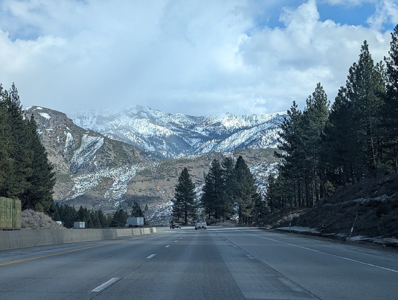 A highway stretches through a forested valley toward snow-capped mountains under a partly cloudy sky.